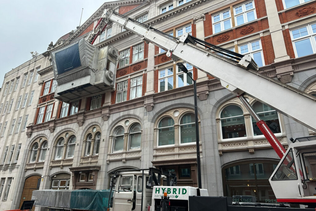 145 Drury Lane, London, during cleaning and restoration by Styles Façade Solutions. A hybrid electric truck-mounted mobile elevating work platform extends its white telescopic boom to raise an enclosed cleaning cabin, wrapped in protective sheeting and branded with scaffold-free façade cleaning signage, against the upper red brick and Portland stone façade. The ground floor arched windows in the granite base are covered with protective plastic film beneath an overcast sky.