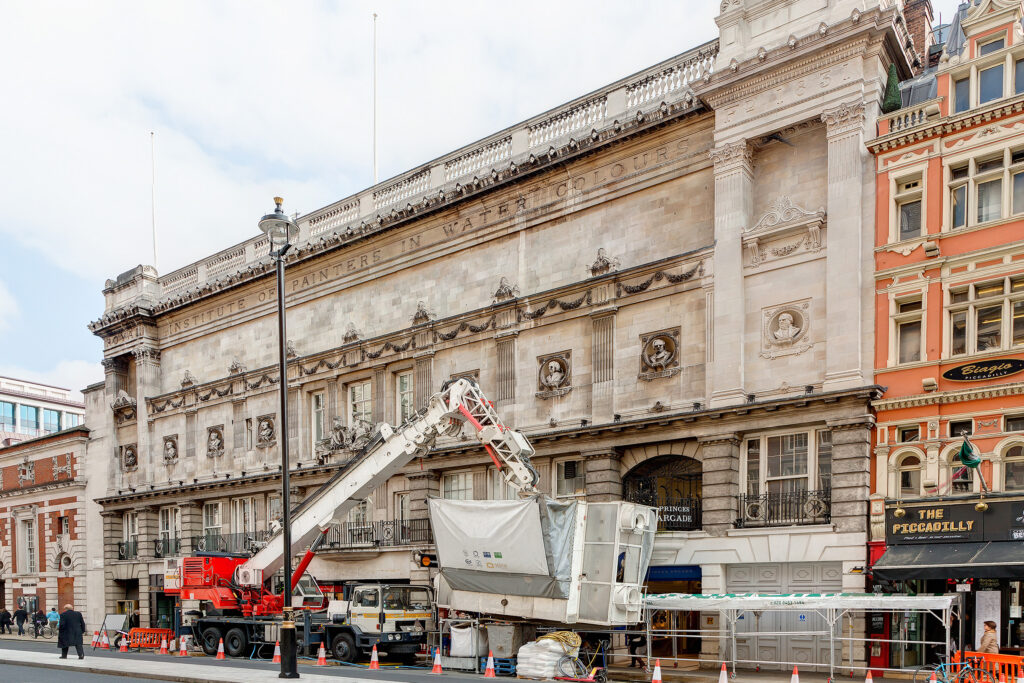 190–195 Piccadilly, London, BAFTA’s headquarters, during Façade Gommage cleaning. A red and white lorry-mounted crane extends its boom to hold an enclosed, sheeted cleaning cabin against the carved Portland stone façade. Traffic cones line the kerb, and a covered walkway protects pedestrians passing the Princes Arcade entrance. Hoses and bulk bags sit on the pavement, far below the sculpted busts and the inscription ‘Royal Institute of Painters in Water Colours’. Central London facade cleaning