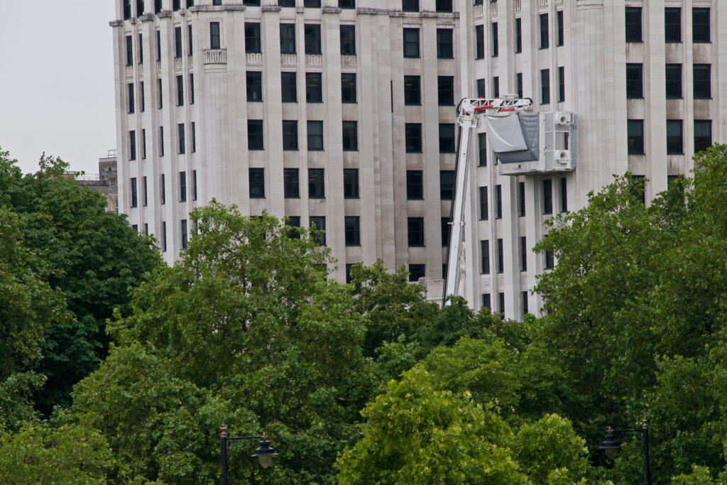Distant view of The Adelphi, London, a Grade II listed Art Deco building, with a truck-mounted specialist cleaning cabin extending high up the tall stone façade during work by Styles Façade Solutions®. The vertical fluted piers and rows of dark-framed windows are partially visible above a dense line of mature green trees. Two ornate lampposts stand in the foreground park below an overcast sky.