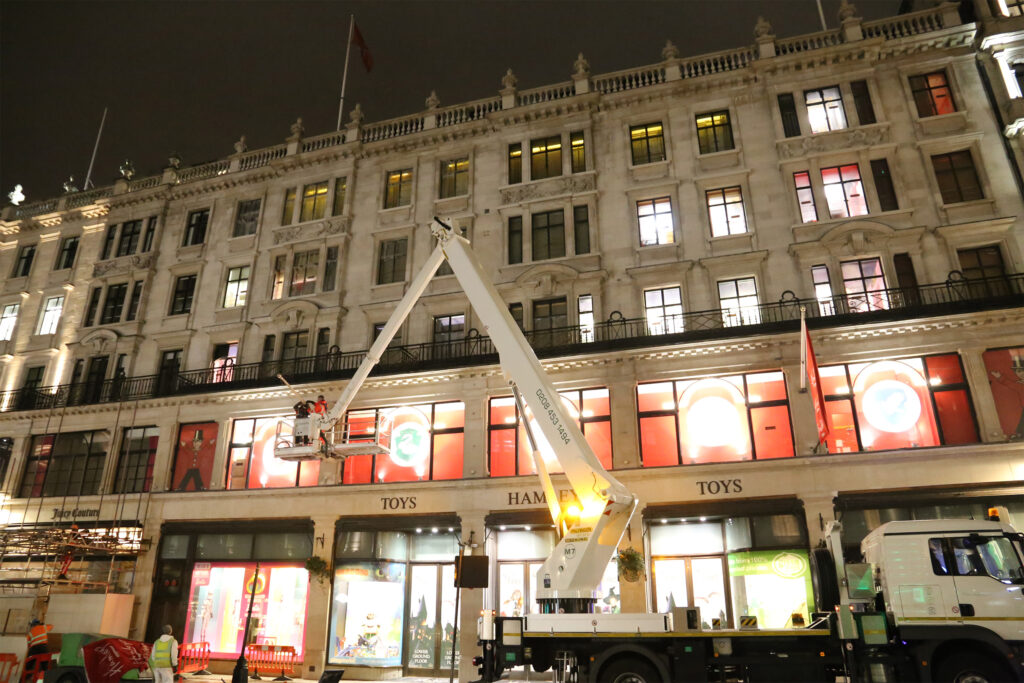 Night-time view of the Hamleys toy store on Regent Street, London, before façade cleaning begins, with the Styles Façade Solutions® team positioned in a mobile elevating work platform, taking down the store's branded flag. The white articulated access truck extends up towards the illuminated first-floor windows, with workers in hi-vis clothing and safety barriers visible on the pavement below.