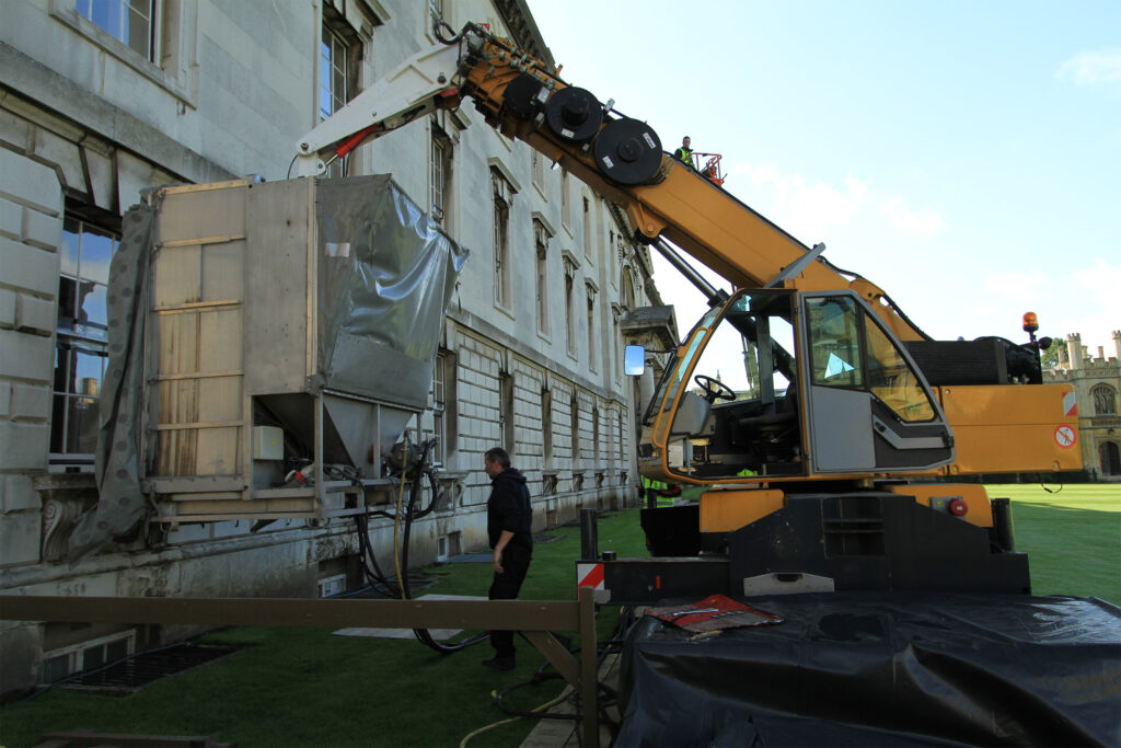 A large yellow cherry-picker crane with an extended arm and mounted equipment carries out façade cleaning work at the Gibbs Building, King's College, Cambridge. An operator works from a platform high on the arm while a worker in high-visibility clothing stands below. A protective silver enclosure and hose system are positioned against the stonework, showing a clear contrast between cleaned and still-stained sections of the façade.