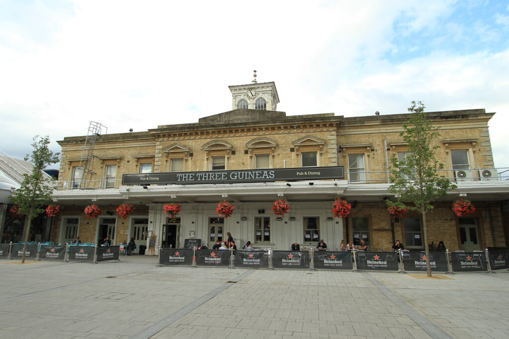 Street-level view of The Three Guineas, a Grade II listed building in Reading, before restoration by Styles Façade Solutions®. The former station building features a central clock tower with weathered stonework, above a pub and dining establishment with a covered terrace decked in hanging red flower baskets. Heineken-branded barriers line the outdoor seating area, with pedestrians seated at tables and a paved square in front.