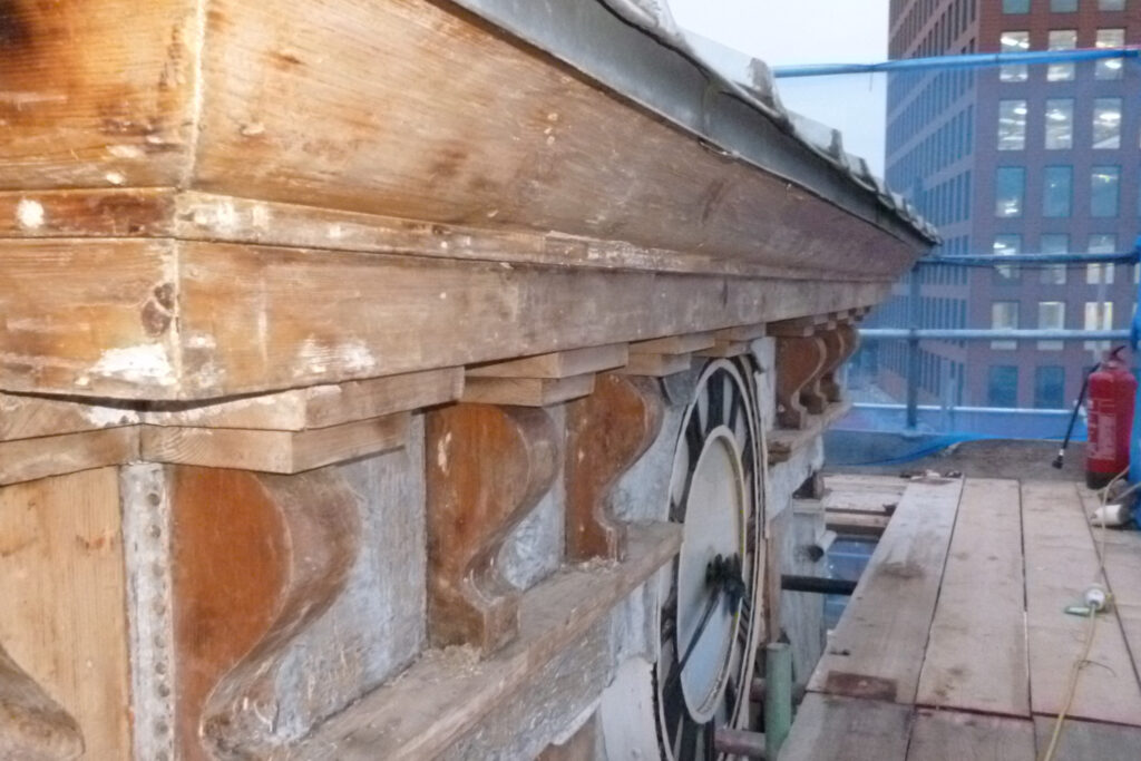 Close-up rooftop view of the clock tower cornice at The Three Guineas, Reading, mid-restoration by Styles Façade Solutions®. The wooden brackets and cornice have been stripped back to bare timber, with the rear of the clock mechanism exposed beside decorative scroll corbels. Scaffolding and a fire extinguisher are visible on the rooftop walkway, with a modern glass-fronted building rising in the background against a pale sky.