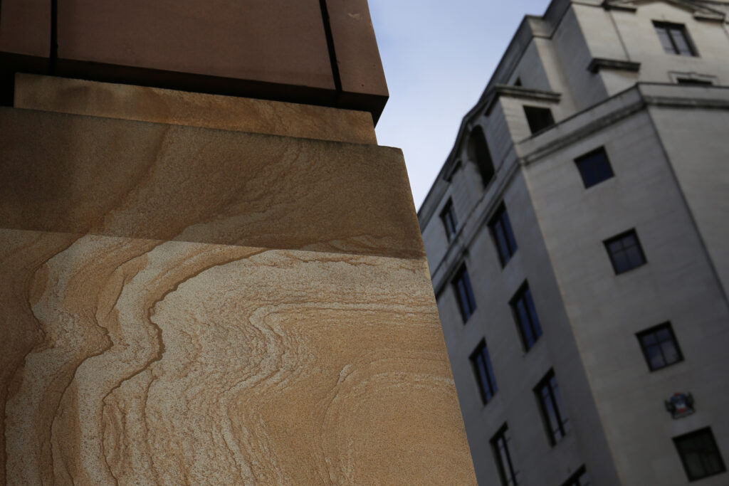 Close-up angled view of a partially cleaned sandstone panel at 1 Poultry, London, during façade cleaning, showing the natural swirling grain and veining of the stone in warm honey tones after treatment with the Façade Gommage cleaning process conducted by Styles Façade Solutions®. Neighbouring grey stone buildings with rows of windows are visible against a pale sky in the background, illustrating the contrast between cleaned and surrounding structures.