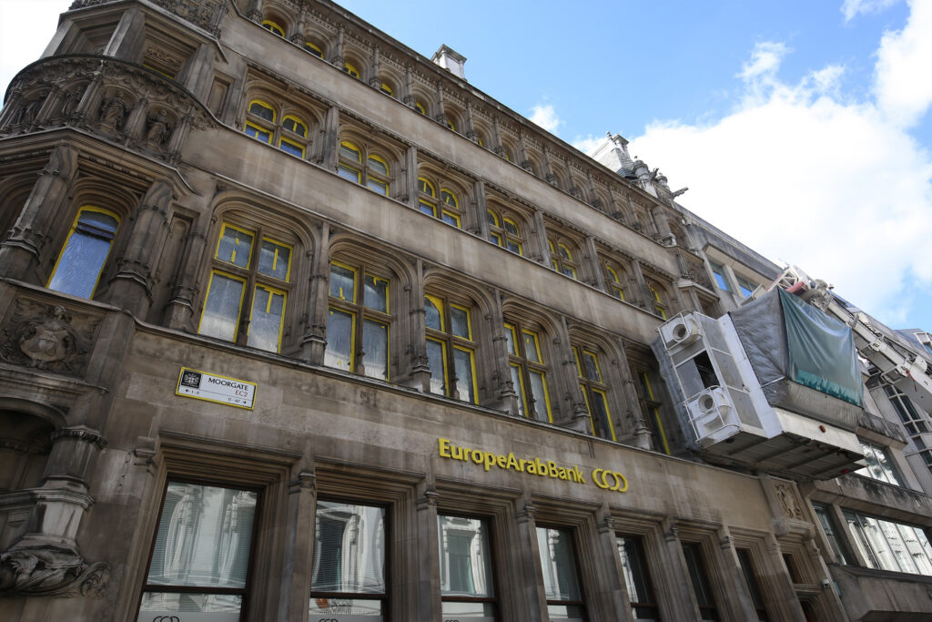 13–15 Moorgate, London, during listed building façade cleaning by Styles Façade Solutions® using the Façade Gommage process. A self-contained cleaning cabin, suspended from a mobile crane, works high on the façade, removing the need for scaffolding. Windows and Europe Arab Bank signage are carefully masked with bright yellow tape and protective sheeting, while the grime-darkened Portland stone, arched windows and statue-filled corner oriel await cleaning beneath a clear blue sky.