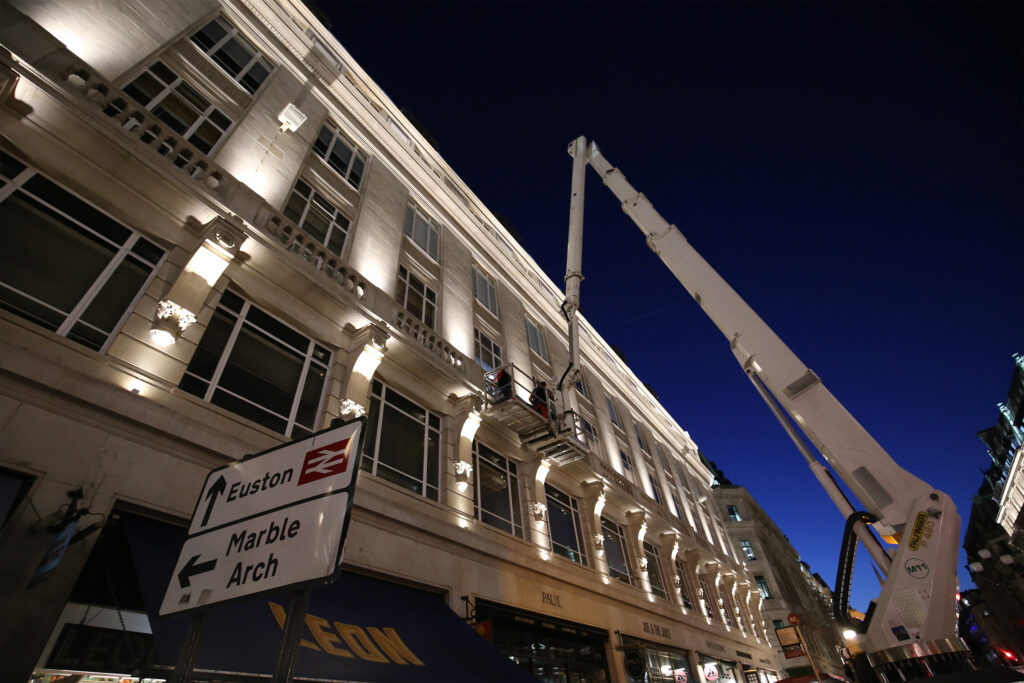 Night-time view of Roxburghe House, London, illuminated against a dark blue sky as workers in a cherry picker platform conduct façade cleaning by Styles Façade Solutions®. The freshly lit stonework and decorative brackets glow warmly, with a street sign for "Euston" and "Marble Arch" in the foreground. Working after hours minimises disruption to ground-floor retailers and the surrounding community below.