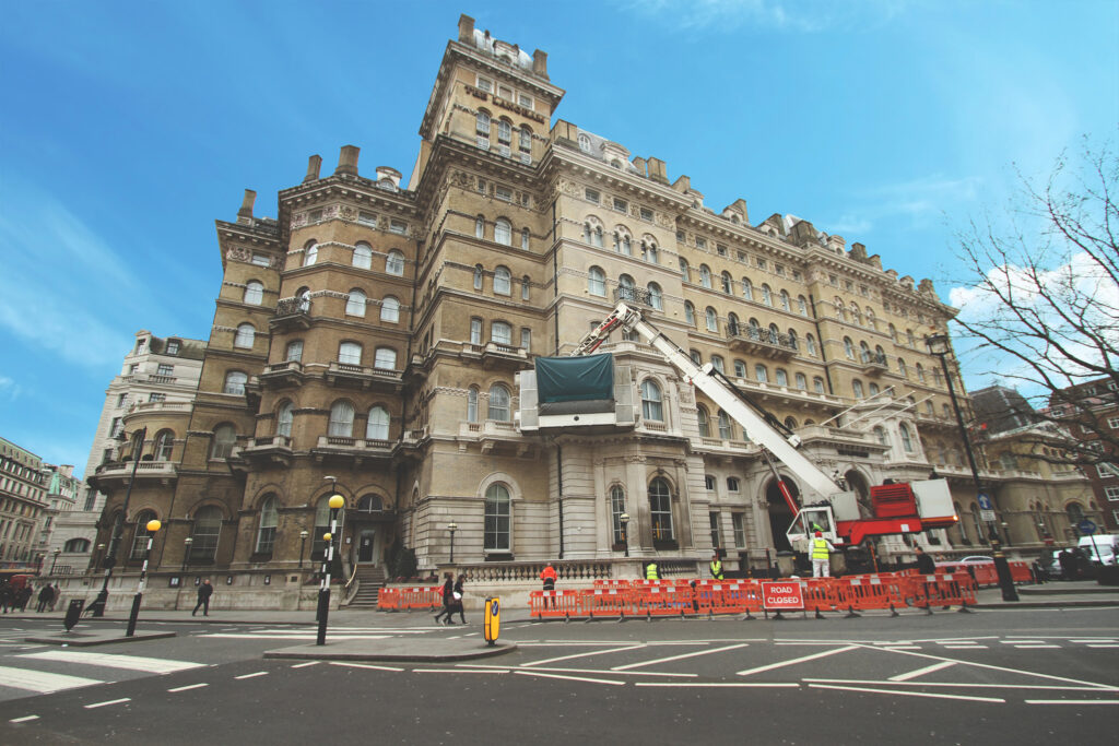Wide-angle view of The Langham Hotel, London, a Grade II listed building, during façade cleaning works. A red and white cherry picker crane with a covered platform reaches up the stone façade as workers in high-visibility clothing operate below, cordoned off by orange barriers and a "ROAD CLOSED" sign. Pedestrians pass by on the pavement beneath a clear blue sky.