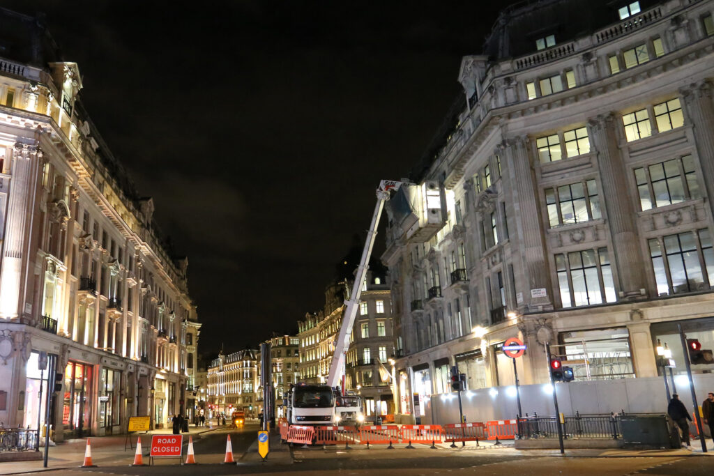 Night-time working view of 253–259 Regent Street (Alitalia House), a Grade II listed building near Oxford Circus, showing a cherry picker access platform raised to upper storeys during Façade Gommage cleaning. Road closure signage, traffic cones and barriers secure the site below, with illuminated shopfronts, Oxford Circus Underground signage and surrounding Regent Street buildings visible under a dark evening sky.