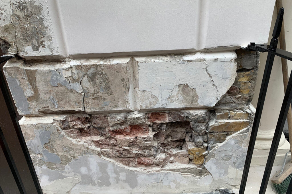 Close-up view of a column base beside the main entrance at 9 Conduit Street, London, during restoration by Styles Façade Solutions®. The image shows extensive render loss exposing the original brickwork beneath, with cracked and delaminating plaster surrounding an area of deep structural damage. Areas of intact painted stonework remain visible above, alongside black wrought-iron railings, illustrating the extent of deterioration requiring repair.