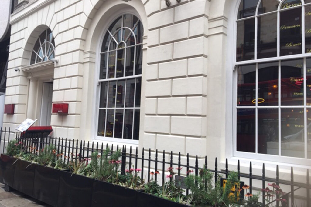Exterior view of 9 Conduit Street, London, before commercial façade restoration by Styles Façade Solutions®. The image shows a period stone building with a cream-coloured ashlar façade, arched sash windows with white timber frames, and decorative stone quoins. Black wrought-iron railings and window boxes with flowering plants line the pavement. A red awning box and menu stand sit near the entrance, with a glazed shopfront reflecting the street beyond.