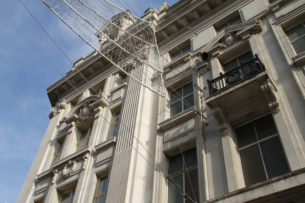 Upper storeys of Grade II listed 266-270 Regent Street, London, during façade cleaning, showing fluted Corinthian pilasters, an ornate pedimented window and a wrought-iron balcony against pale, evenly cleaned stonework. Overhead decorative lighting rigging and cabling cross the façade, with clear blue sky beyond. The crisp, bright tone of the stone highlights the results already achieved through the ongoing heritage building façade cleaning works.