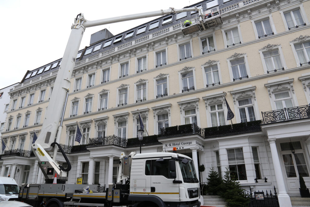 Cream-painted Victorian stucco terrace at 1-15 Templeton Place, London, home to the K+K Hotel George, photographed before façade repair and restoration by Styles Façade Solutions®. A white Palfinger cherry picker mounted on a MAN truck extends to roof level, where a surveyor in a harness inspects the parapet and mansard slate roofline. Ornate window pediments, wrought-iron balconies, and ground-floor columned entrance porch are visible along the multi-storey frontage.