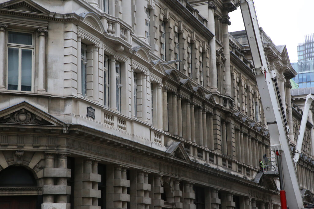 Close-up angled view of 1-5 London Wall, London, during façade cleaning, showing the ornate classical stonework appearing bright and freshly cleaned, with decorative columns, balustrades, and a "London Wall" street sign. Protective covering is fitted to windows, while a white crane with an access cage carrying a worker in hi-vis clothing operates near the building's upper storeys against a pale sky.