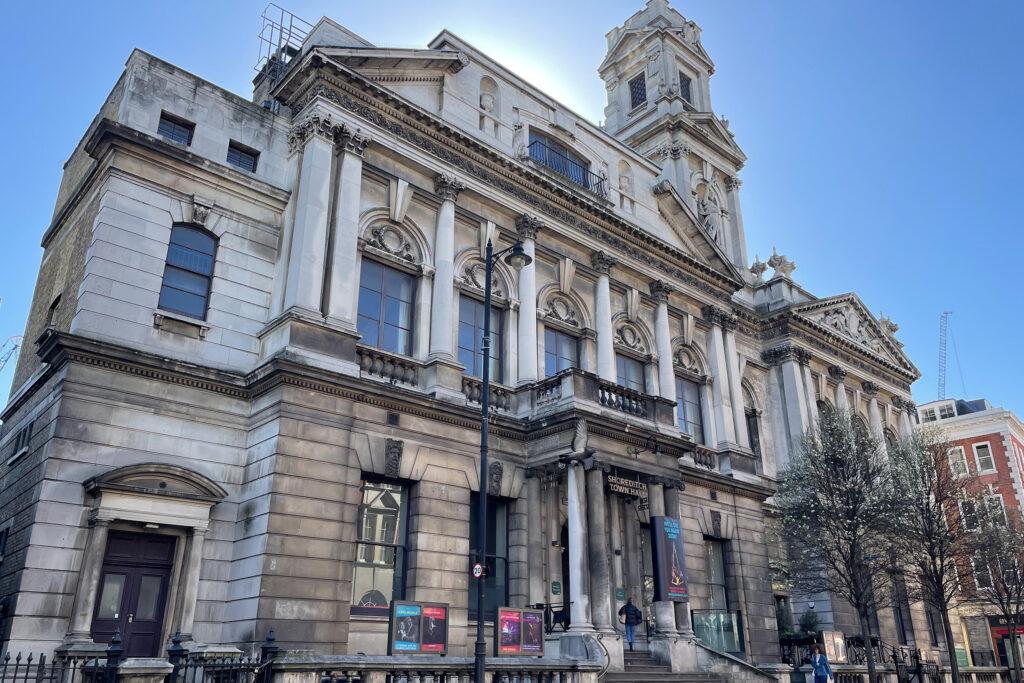 Street-level view of Shoreditch Town Hall, London, a Grade II listed Baroque-style building with ornate stone façade, columns, pediments, and a prominent clock tower, photographed in bright sunlight before restoration work begins. The weathered stonework shows decades of accumulated grime and staining. Entrance banners and posters are visible near the main doors, with a pedestrian walking past and bare trees lining the street in front.