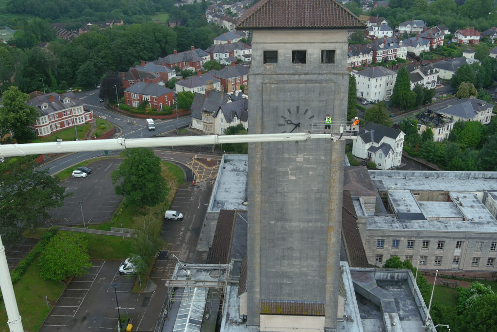 Aerial view of Newport Civic Centre's Grade II* listed clock tower, showing its stone façade and clock face up close. Two workers in high-vis clothing stand on a cherry picker platform beside the tower, conducting a façade survey. Below, the surrounding rooftops, streets, parked cars, and residential terraces of Newport are visible, with mature green trees framing the scene on a overcast day.
