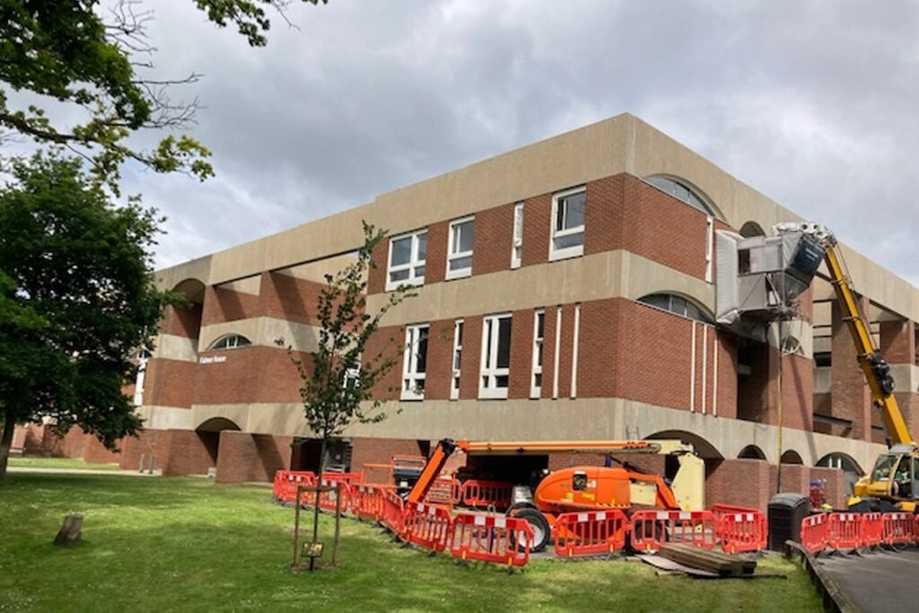 Falmer House, University of Sussex, during façade cleaning by Styles Façade Solutions® using the Façade Gommage process. A yellow crane extends an enclosed access platform toward the upper brick and concrete façade, while an orange access platform waits on the grass below. Red and white safety barriers cordon off the equipment area, with the building's distinctive banded brickwork and concrete detailing visible under a cloudy sky.