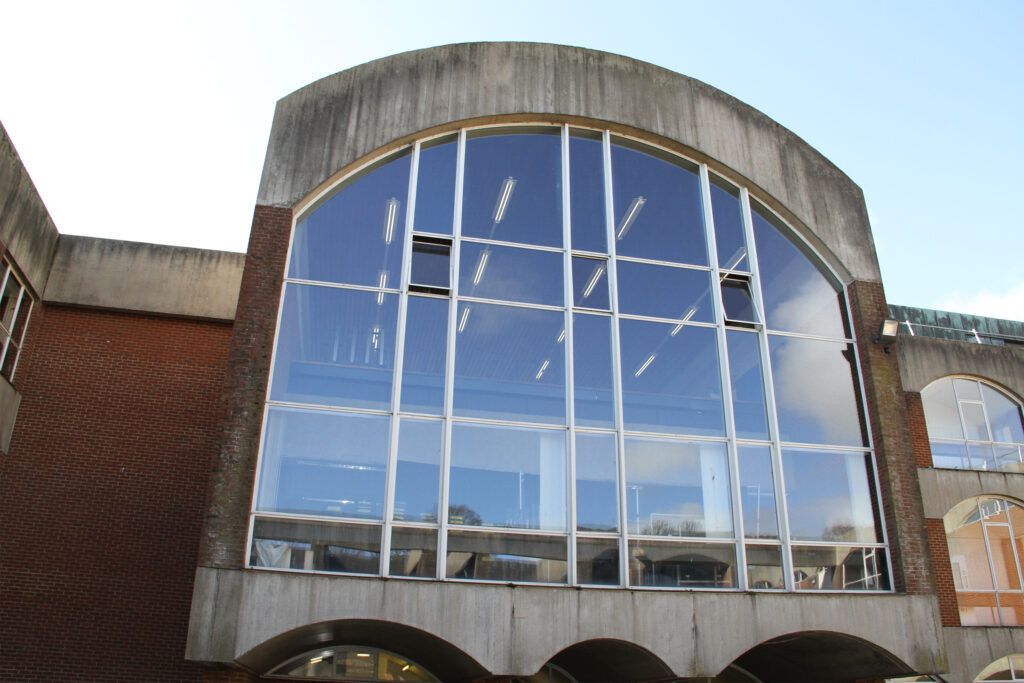 Styles Façade Solutions® is set to clean Falmer House, University of Sussex, using the Façade Gommage process. This "before" image shows the building's weathered concrete and brick exterior, marked by heavy staining and algae growth around the distinctive curved glass window wall, ahead of restoration work.