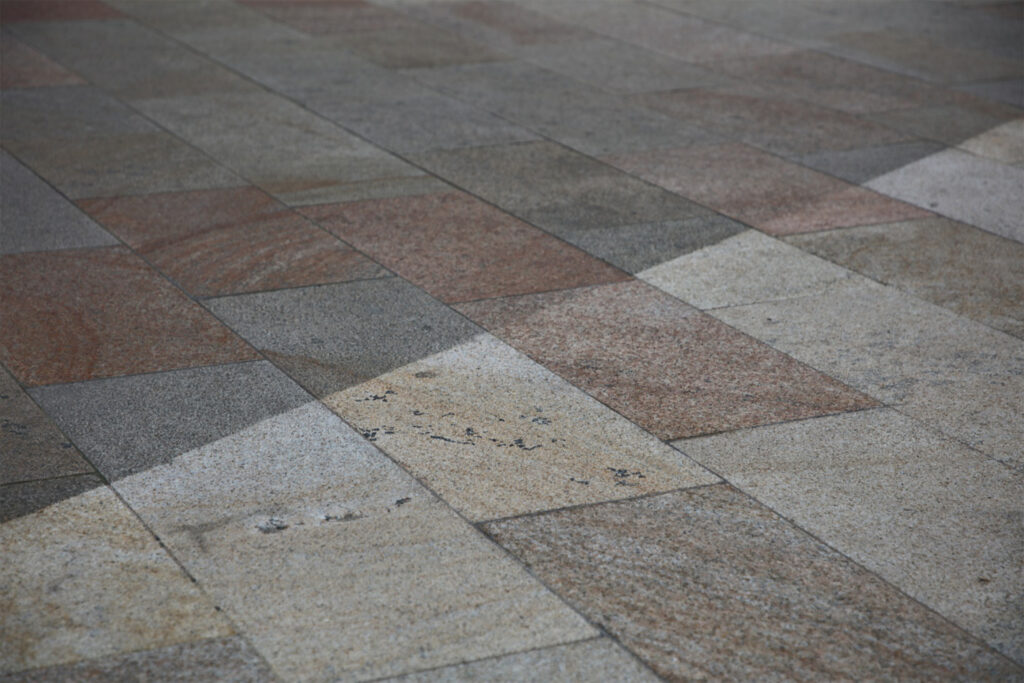 Styles Façade Solutions® carries out public realm cleaning on granite footpath paving. The close-up image shows a clear contrast between darker, soiled stone sections and lighter, cleaned areas, demonstrating the effectiveness of the cleaning process in restoring the pavement's natural colour and finish.