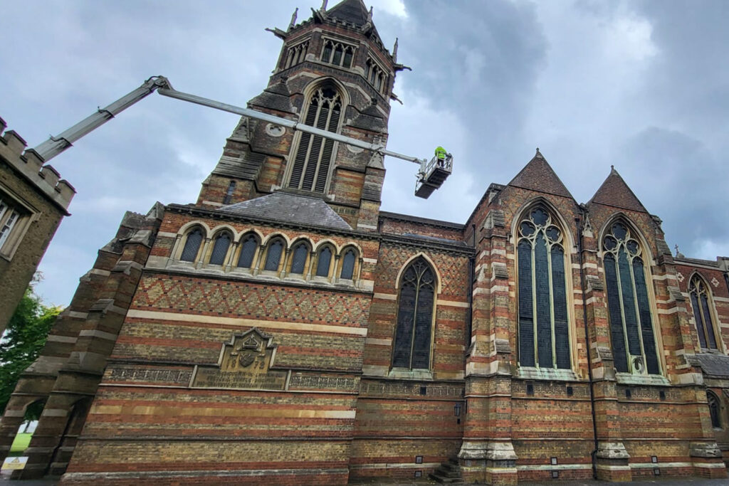 The Grade I listed Rugby School Chapel, Rugby, during a façade survey by Styles Façade Solutions®. A MEWP extends high up the polychrome brick tower, with a technician in high-visibility gear inspecting stonework near the louvred belfry windows. The chapel's Gothic Revival architecture features patterned brickwork, arched windows and a datestone reading 1872, set beneath a cloudy sky with a neighbouring building visible to the left.