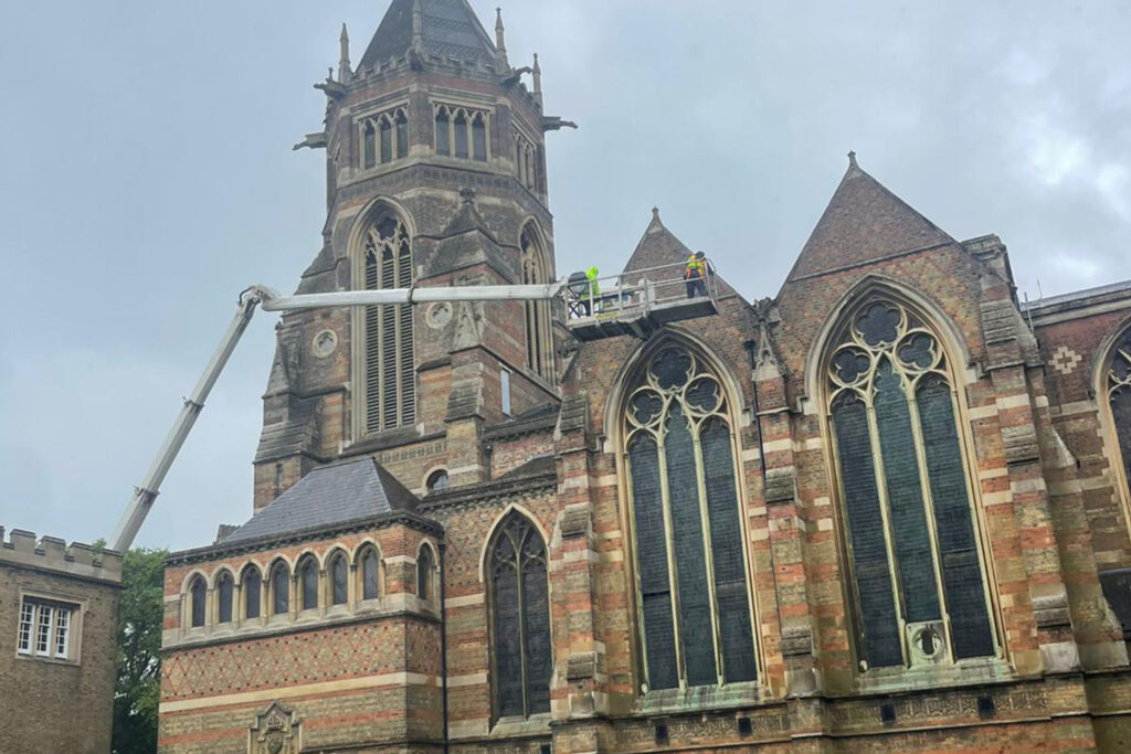 Styles Façade Solutions® conducts a façade survey at the Grade I listed Rugby School Chapel in Rugby. A MEWP platform carries two technicians across the roofline to inspect the tower and gable, highlighting the chapel's ornate Gothic Revival brickwork and tracery windows.