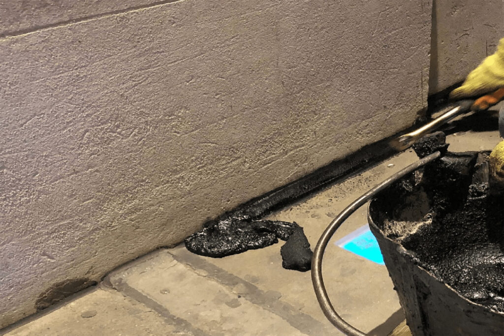 Close-up of a technician in yellow gloves applying hot black bituminous sealant along the base joint where a stone wall meets the pavement, using a torch and brush. A blue flame from the heating torch is visible, softening the material for application. Wet sealant coats the junction, ensuring a weatherproof seal between the masonry and ground surface below.