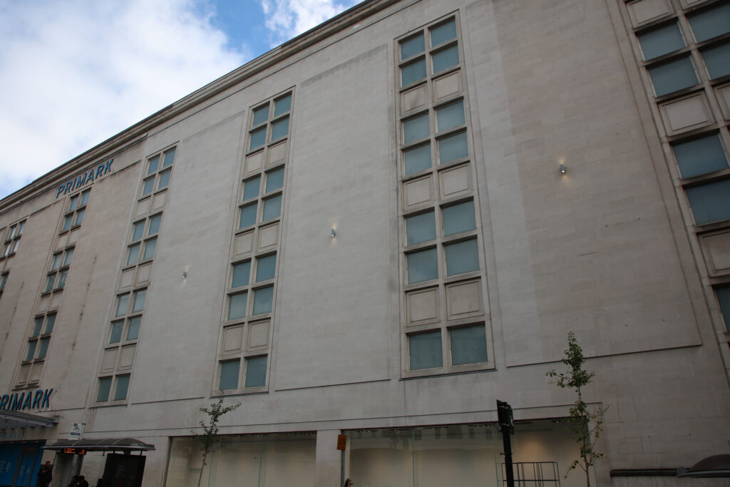 A daytime view of the Portland stone façade at 1-27 Horsefair, Bristol, home to Primark, showing the results of overnight cleaning by Styles Façade Solutions®. A clear contrast is visible between freshly cleaned, lighter stone sections and adjacent areas still awaiting treatment, with tall grid-patterned windows running up the building. Blue skies and scattered clouds provide a bright backdrop to the visible progress.