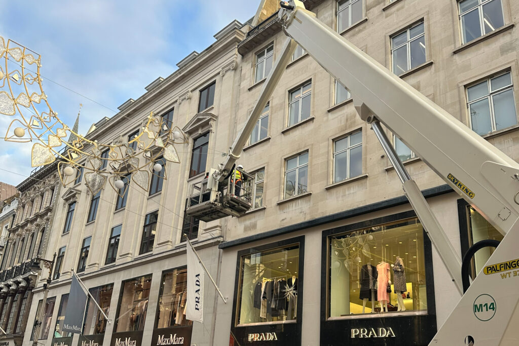 A surveyor in high-visibility clothing and a hard hat inspects the upper stonework of 16-18 Old Bond Street, London, from a cherry-picker platform during a commercial façade survey by Styles Façade Solutions®. The extended access arm reaches the third-storey windows of the Portland stone building, with the Prada and MaxMara shopfronts visible below, alongside decorative overhead street lighting and a flag reading "Prada."