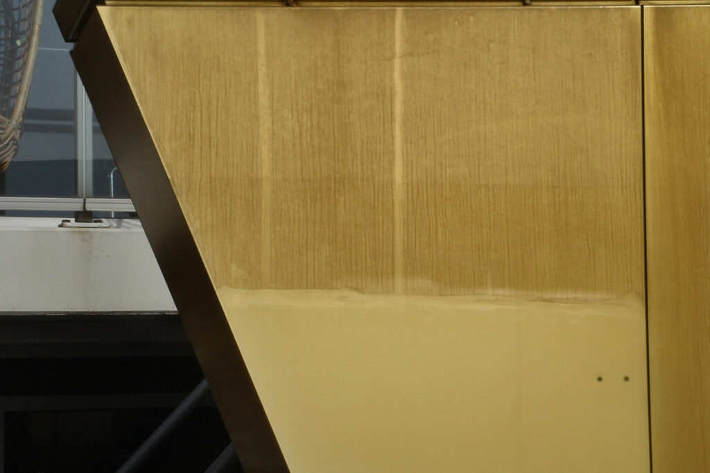 Styles Façade Solutions® demonstrates the striking transformation of a gold coloured anodised aluminium panel, cleaned diagonally to reveal a bright, glossy finish beside the original weathered surface. The clear before-and-after contrast highlights the effectiveness of the restoration process and the panel's renewed golden appearance.