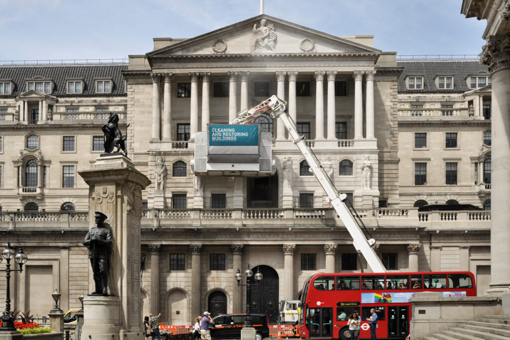 The Portland stone façade of the Bank of England, London, photographed during cleaning works. A Styles Façade Solutions (formerly Thomann-Hanry®) Façade Gommage cleaning unit, mounted on an extended hydraulic boom arm, is positioned against the neoclassical elevation. A banner on the unit reads "Cleaning and Restoring Buildings." London street life continues below, with a red double-decker bus, pedestrians, and a bronze monument visible in the foreground.