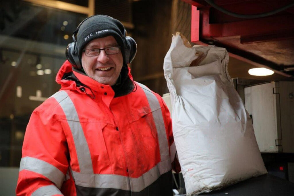 A smiling Styles Façade Solutions (formerly Thomann-Hanry®) operative wearing a red high-visibility jacket, black beanie hat and ear defenders, holding a large paper sack of Façade Gommage cleaning powder beside the cleaning vehicle. The team member appears to be loading materials into the specialist cleaning unit in preparation for façade treatment works. Industrial equipment and a building interior are visible in the background.