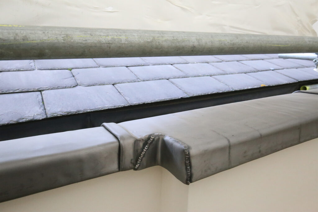 Close-up of newly repaired lead flashing along a roof edge, completed by Styles Façade Solutions. The lead parapet gutter features a neatly welded joint with visible burned lead seams, sitting beneath rows of grey slate roof tiles. A scaffold pole runs along the top, with protective sheeting visible in the background and a cream-rendered wall below the lead detail.