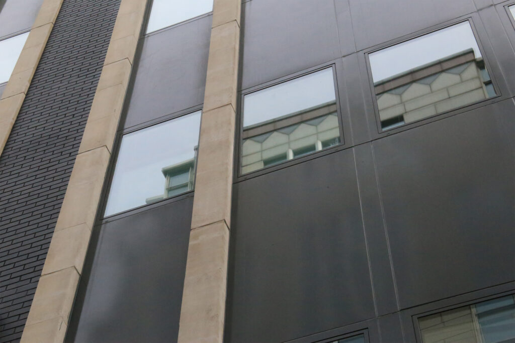 An upward view of a modern building façade featuring dark anodised aluminium window panels set between pale stone piers, with dark brick cladding to the left. Reflections of neighbouring rooftops appear in the glazing. The aluminium panels show visible wear and discolouration prior to restoration works being undertaken by Styles Façade Solutions to refurbish the metal cladding.