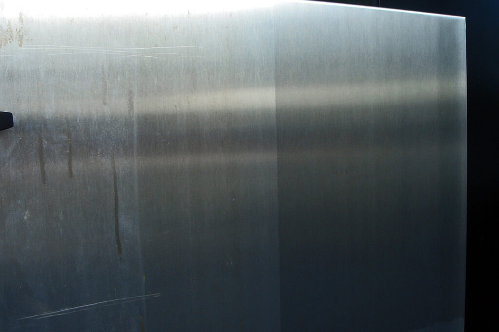 Styles Façade Solutions® mid-restoration of stainless steel panels, capturing heavy staining, streaking, and scratches across its dulled surface. The weathered condition highlights the need for professional cleaning and restoration to return the panel to its original brushed metal finish.