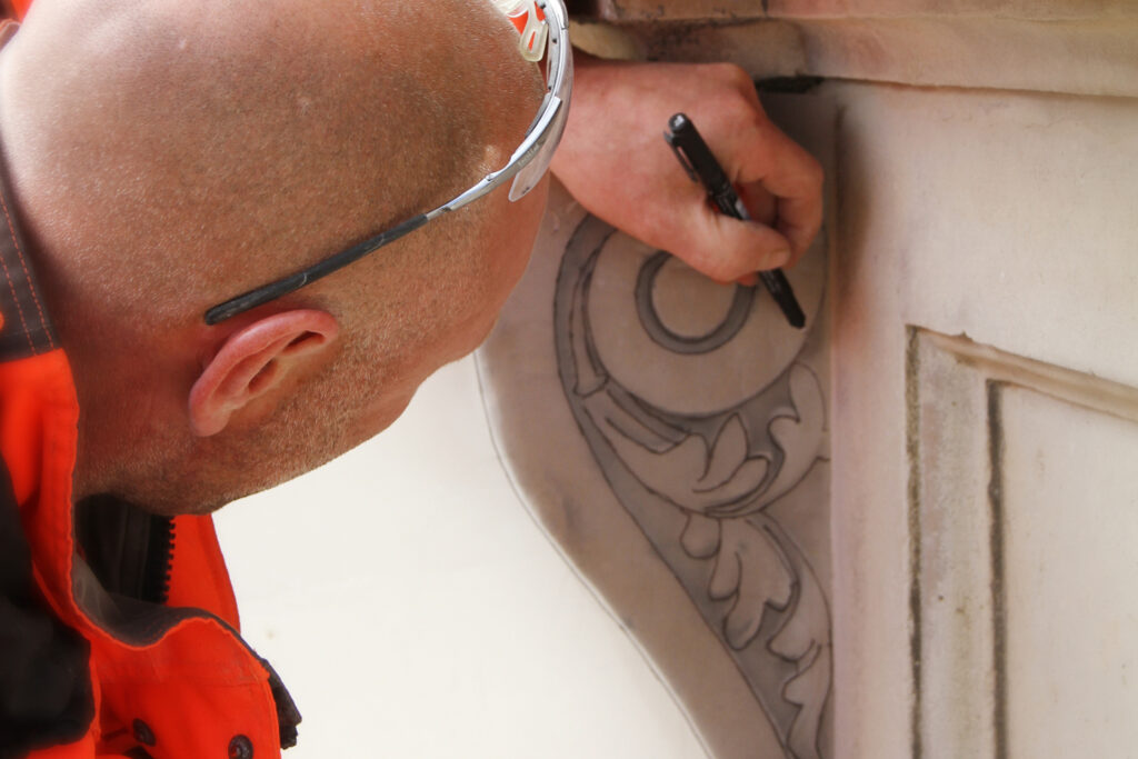 A stonemason wearing safety glasses and an orange hi-vis jacket leans in close to trace a decorative scroll and acanthus leaf motif onto tracing paper held against a stone façade detail at Gibb's Building, King's College, Cambridge. Using a black marker, he carefully records the ornamental design during restoration works, preparing to recreate the carving as a replacement piece back in the workshop.