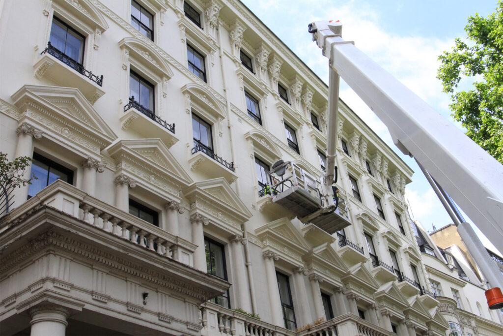 Styles Façade Solutions® deploy their specialist truck-mounted boom lift to conduct a detailed mid-level façade survey on an ornate white stucco Victorian property in central London. Two operatives in the elevated platform basket carry out close-up inspection of the building's richly decorated cornices, pilasters, and elaborate window surrounds.