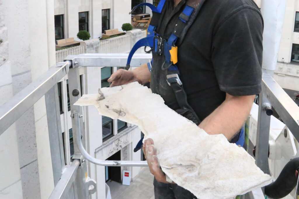 A Styles Façade Solutions survey operative wearing a full safety harness with blue straps stands in an elevated platform basket, holding a large fragment of delaminated stone or render material removed from a building façade during a high-level condition survey. The substantial piece of failed façade material, displaying visible cracking, delamination, and soiling, clearly demonstrates the structural deterioration identified during the scaffolding-free survey operation at high level.