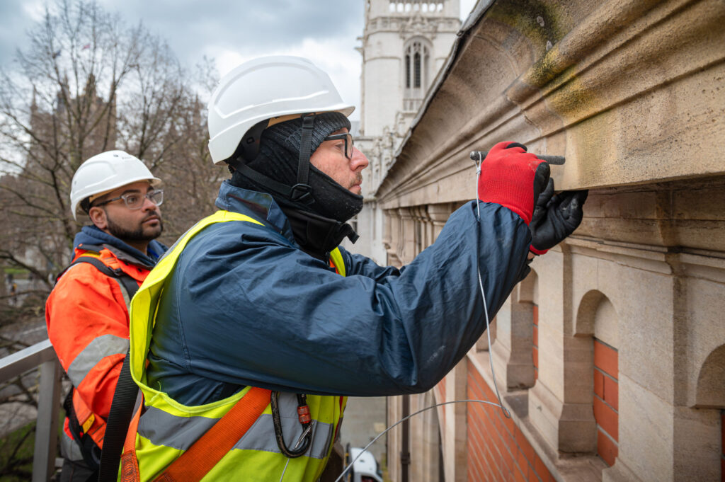 A surveyor in hi-vis clothing and white safety helmet inspect a stone balustrade and cornice on a high-level building ledge, with a Gothic church tower visible in the background. One worker examines a wire fixing closely while his colleague observes. Moss and weathering are visible on the stonework. This careful inspection forms part of a façade survey carried out by the skilled team at Styles Façade Solutions (formerly Thomann-Hanry®). commercial facade surveys