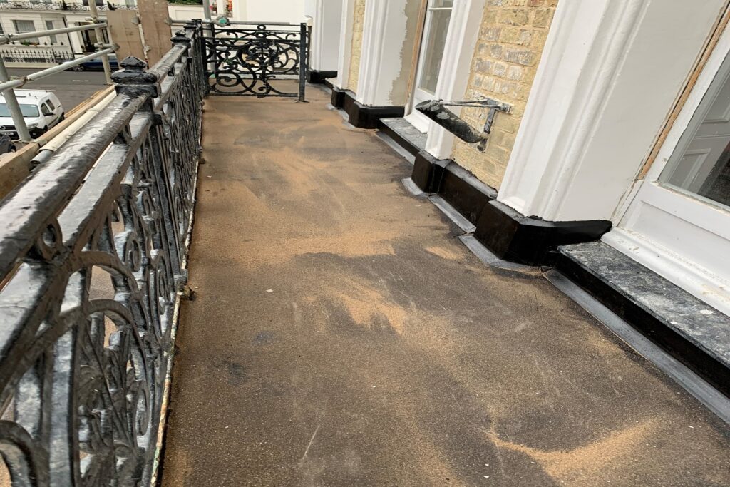 View along a newly asphalted balcony walkway on a period townhouse, bordered by an ornate black wrought-iron railing. Freshly applied felt roofing covers the deck, with new lead flashing detailing around the base of white-painted window surrounds and yellow brickwork. Scaffolding and a street of Georgian-style terraced houses are visible in the background under an overcast sky.