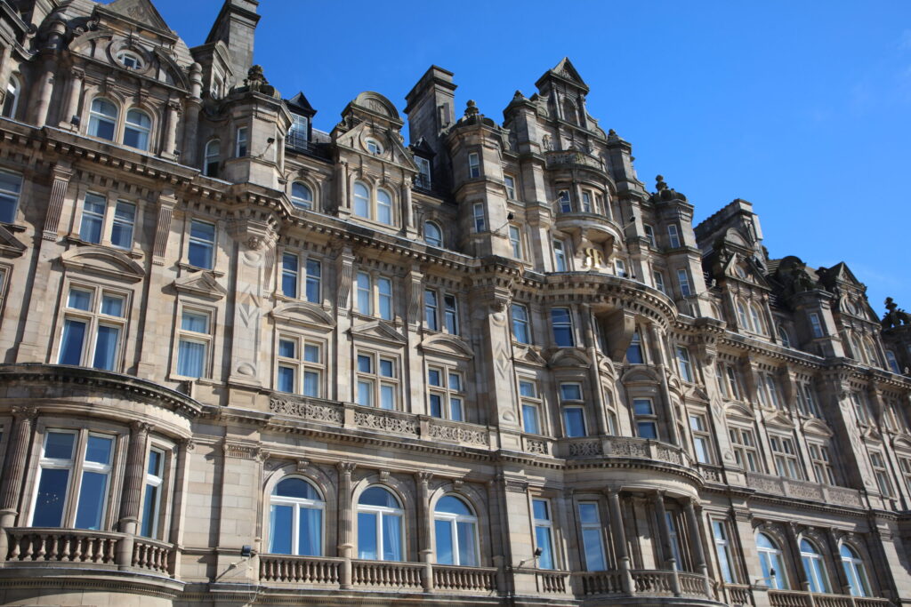 Façade of The Balmoral Hotel featuring ornate Victorian stone architecture, with decorative columns, arched windows, balconies, turrets, and intricate carvings. The historic building is illuminated by sunlight beneath a clear blue sky.