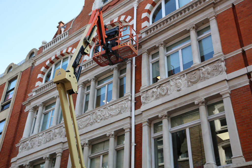 Styles Façade Solutions (formerly Thomann-Hanry®) carrying out window repair and redecoration works at 14–15 Conduit Street, London, using a MEWP (Mobile Elevating Work Platform). The brilliantly restored Portland stone pilasters, arched window surrounds and intricately carved decorative frieze of mascarons and foliate scrollwork following a Façade Gommage cleaning.