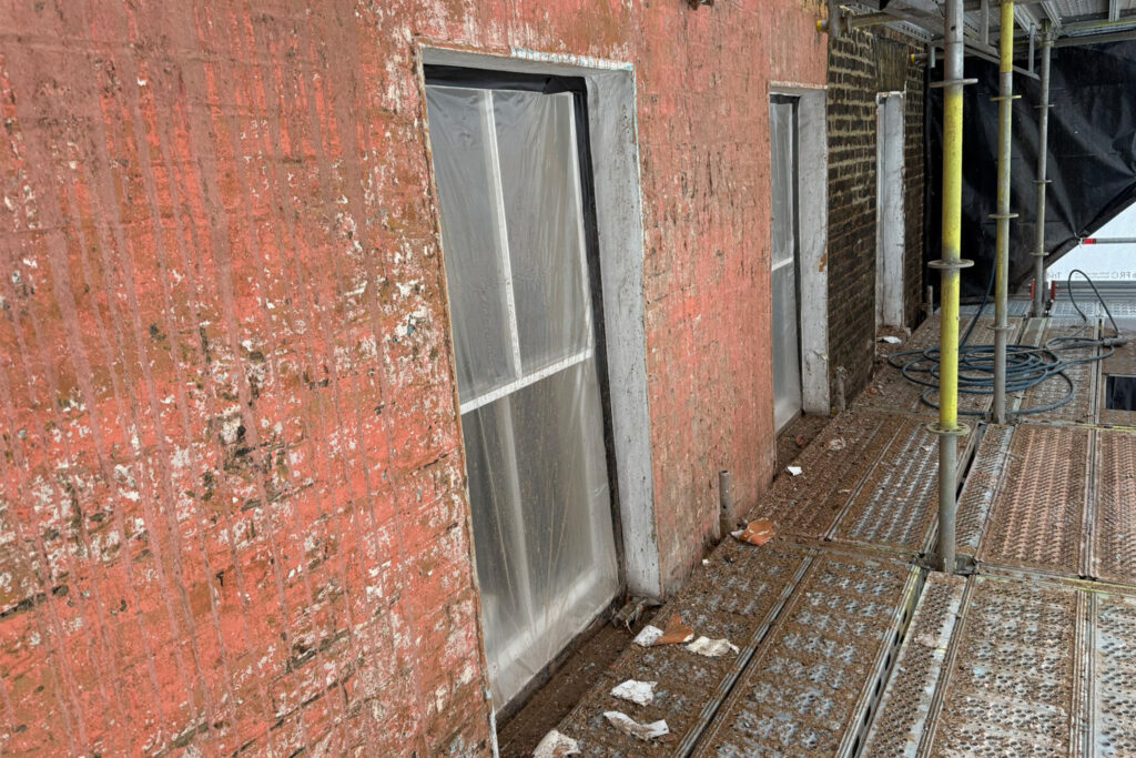 A close-up view of the façade of 46 Carnaby Street, London, photographed during restoration works from scaffolding level. The brickwork is mid-process, with layers of light blue and red paint partially stripped to reveal the underlying brick beneath. Sash windows are protected with clear plastic sheeting. Debris and paint flakes are visible on the scaffolding platform, with yellow scaffold poles and equipment visible to the right.