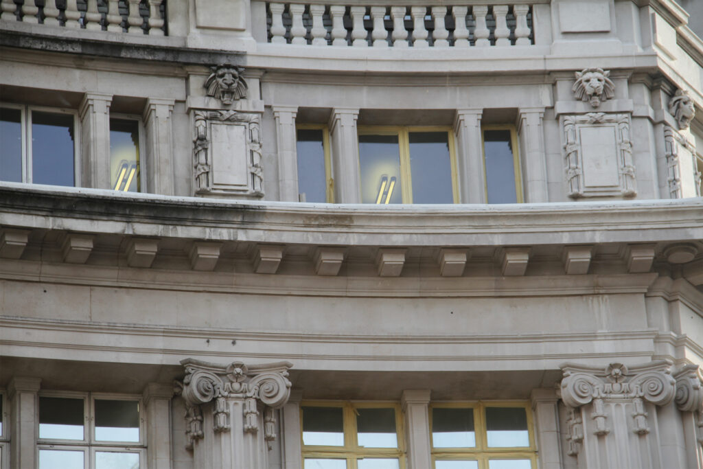 Styles Façade Solutions® carrying out Façade Gommage cleaning on Spirella House, Regent Street. The close-up reveals a sharp contrast between soiled and restored stonework around carved lion heads and Ionic capitals, showcasing the precision of the conservation grade cleaning process on ornate detailing.