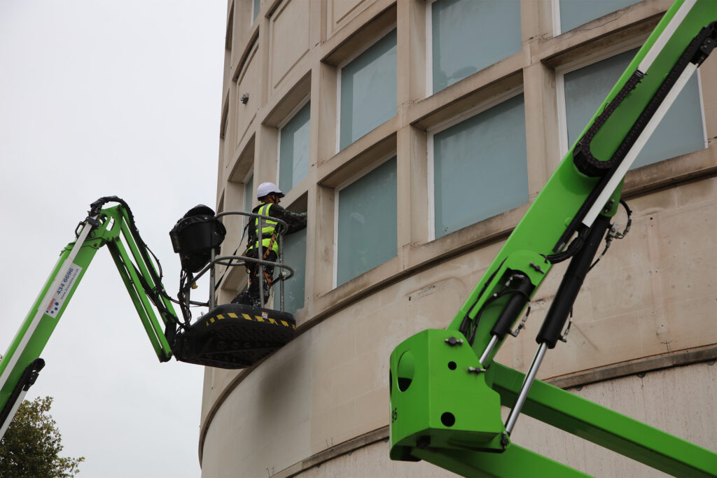 Styles Façade Solutions® carrying out scaffold-free window painting at Primark, Broadmead, Bristol. An operative works from a MEWP, painting frames set within the curved stone façade, demonstrating an efficient access method for maintenance works without the need for full scaffolding.