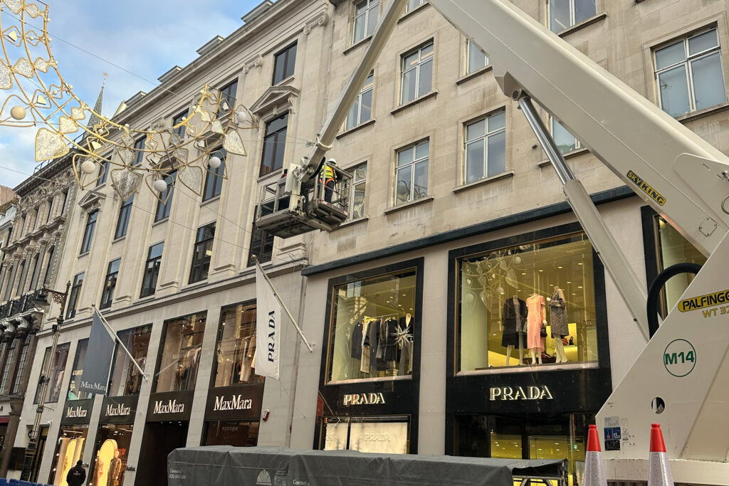 Styles Façade Solutions® conducting a façade condition survey of Prada, Bond Street. An operative on a cherry-picker inspects the soiled Portland stone upper storeys, assessing the building's condition above the Prada storefront ahead of possible cleaning and restoration works.