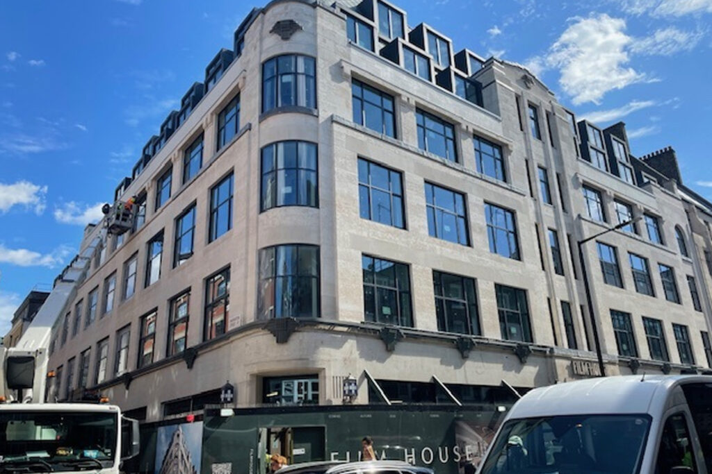 Styles Façade Solutions (formerly Thomann-Hanry®) mid-clean at Film House, 142 Wardour Street, London. A hydraulic boom lift positions an operative at upper storey level as the Façade Gommage process progressively transforms the Portland stone façade, with the majority of cleaning already visibly completed.