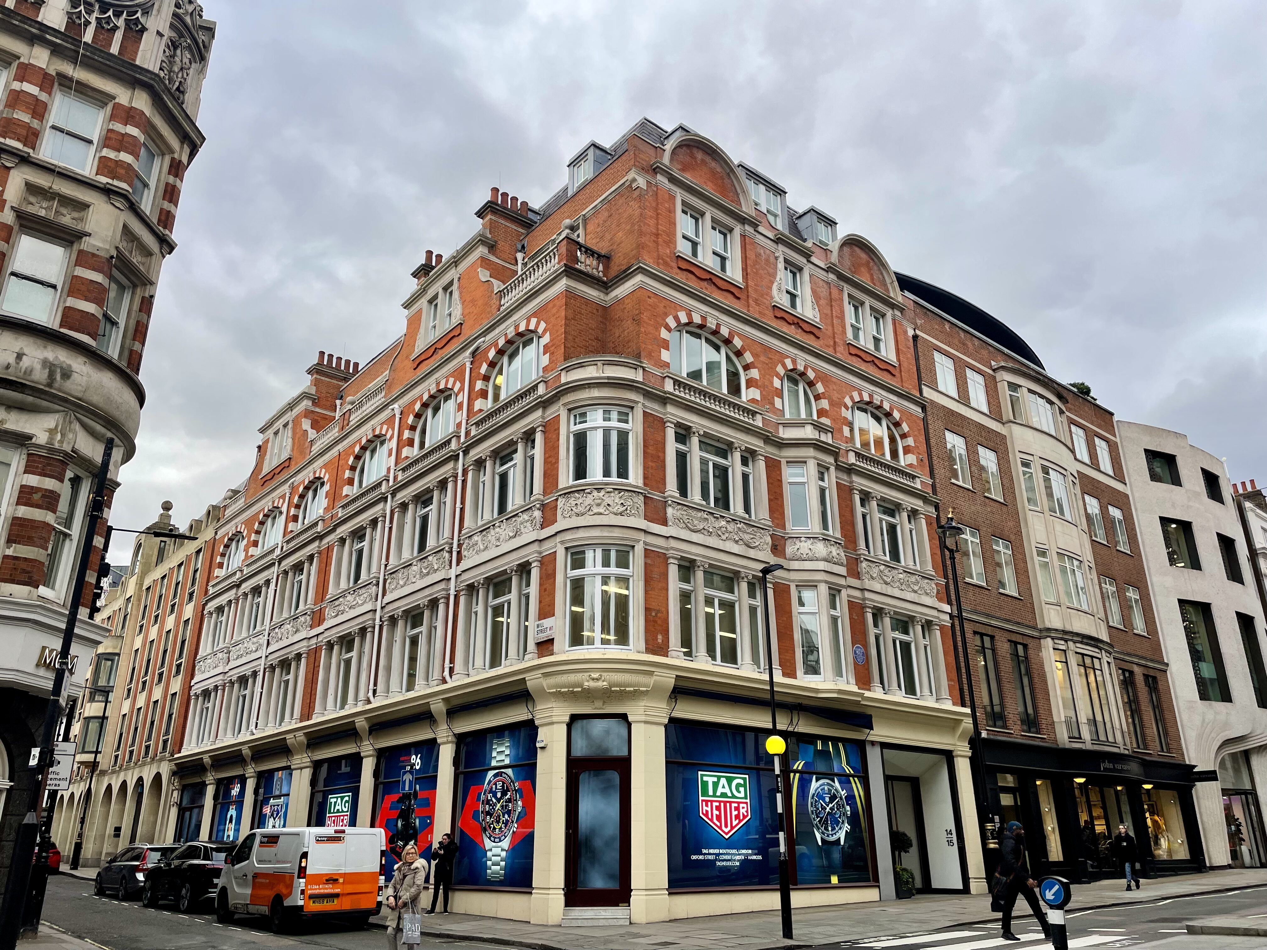 Corner view of a Victorian red brick and Portland stone building at 14-15 Conduit Street, London, with arched windows, decorative stone banding, and ornamental carved panels. Pedestrians and parked vehicles line the street. The crisp brickwork and stonework reflect recent cleaning and redecoration by Styles Façade Solutions (formerly Thomann-Hanry®). office facade cleaning
