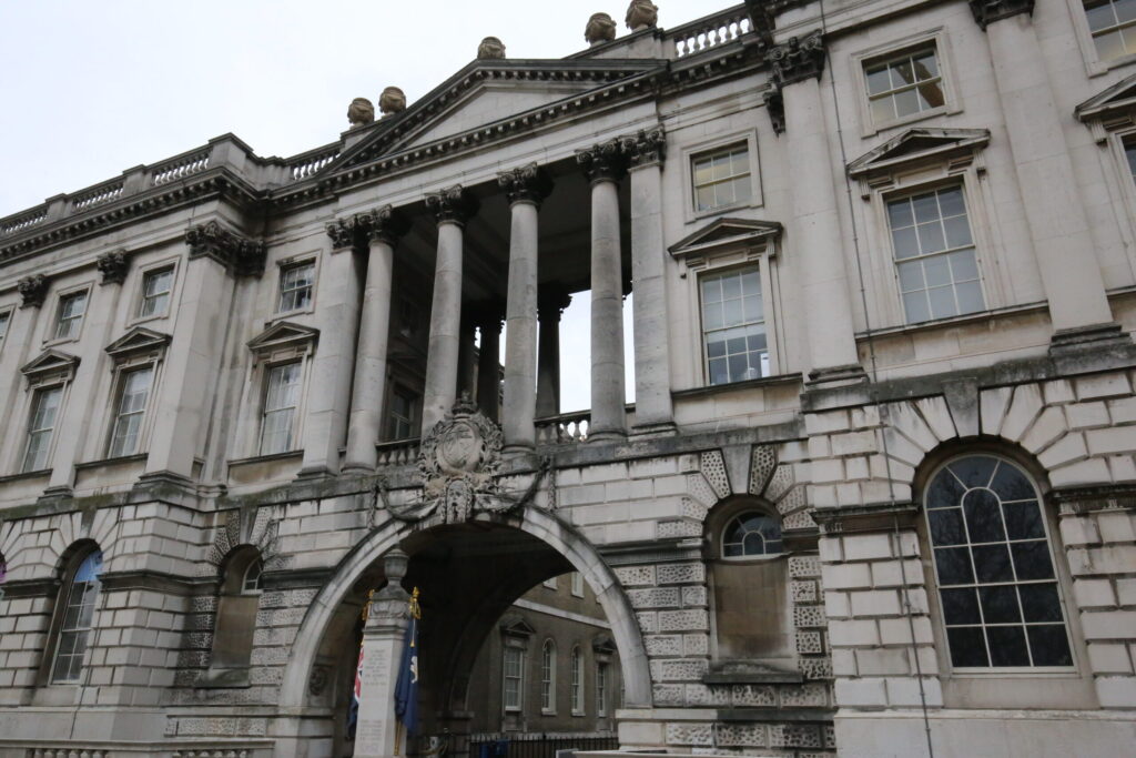 Weathered Portland stone façade at Somerset House, London, featuring a grand Corinthian colonnade, carved heraldic crest with crown and swags above an archway, and decorative roofline urns. Streaks of dark biological staining and grime are visible across the stonework, particularly around window surrounds and the lower archway. This soiled condition illustrates the building's appearance prior to the Façade Gommage cleaning process conducted by Styles Façade Solutions (formerly Thomann-Hanry®).