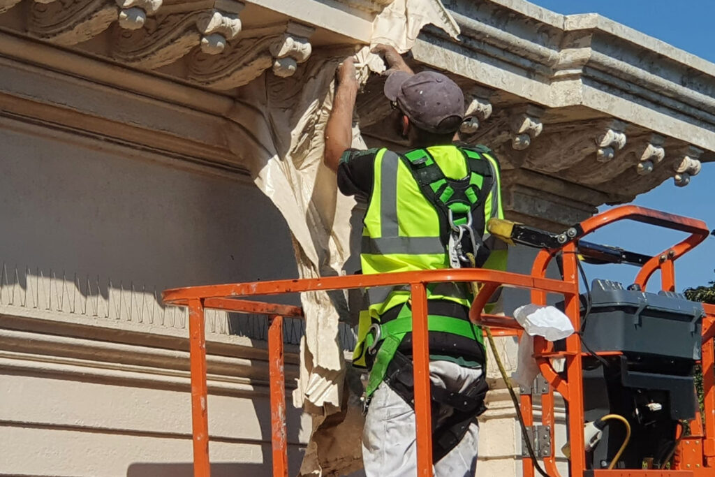 A Styles Façade Solutions operator, wearing a high-visibility vest and safety harness, stands in an orange cherry picker basket while stripping loose, peeling paint from ornate stone corbels beneath a decorative cornice on a heritage building. Large sheets of old paint are pulled away by hand, revealing the carved detailing underneath, with bird-deterrent spikes visible along the ledge below.
