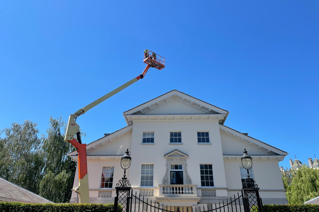 A large white Georgian-style heritage building with a triangular pediment roofline, sash windows, and ornate iron gates is photographed against a clear blue sky. A cherry picker (mobile work elevating platform) extends high above the roofline, positioning two operators from Styles Façade Solutions in the basket, preparing to begin exterior façade painting and restoration work on the upper sections of the property's classical elevation.