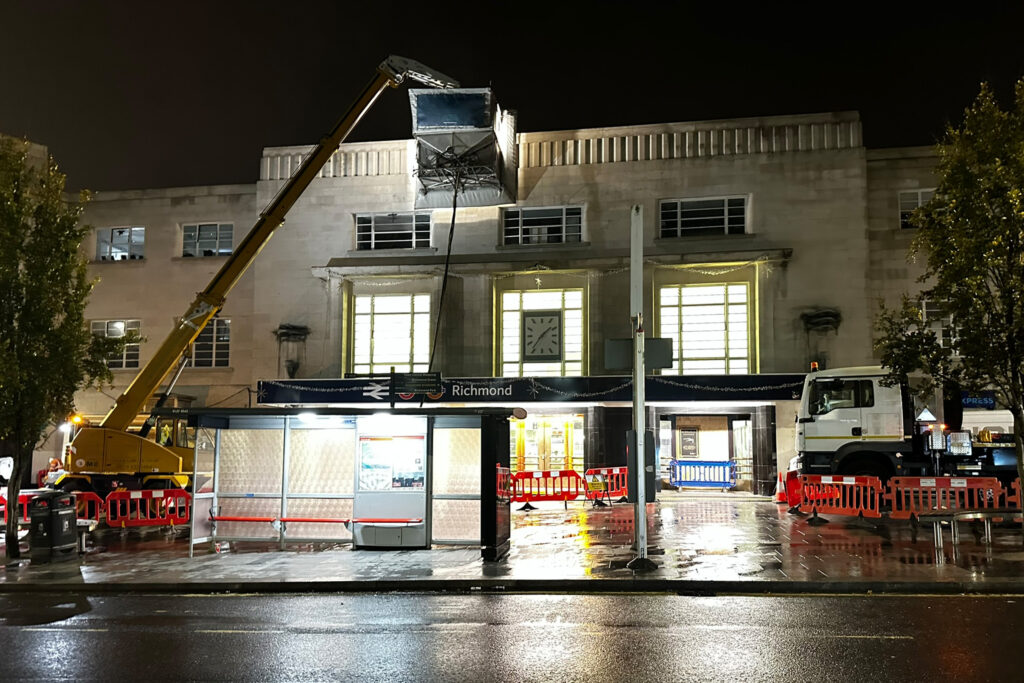 The Art Deco Portland stone façade of Richmond Station, London, during night-time Façade Gommage cleaning works by Styles Façade Solutions (formerly Thomann-Hanry®). The specialist cleaning cabin, suspended from a yellow hydraulic crane arm, is positioned at upper parapet level against the illuminated station frontage. Orange safety barriers cordon the wet forecourt, with the crane support vehicle visible to the right. The station clock, Richmond signage and Christmas lights are visible on the façade.